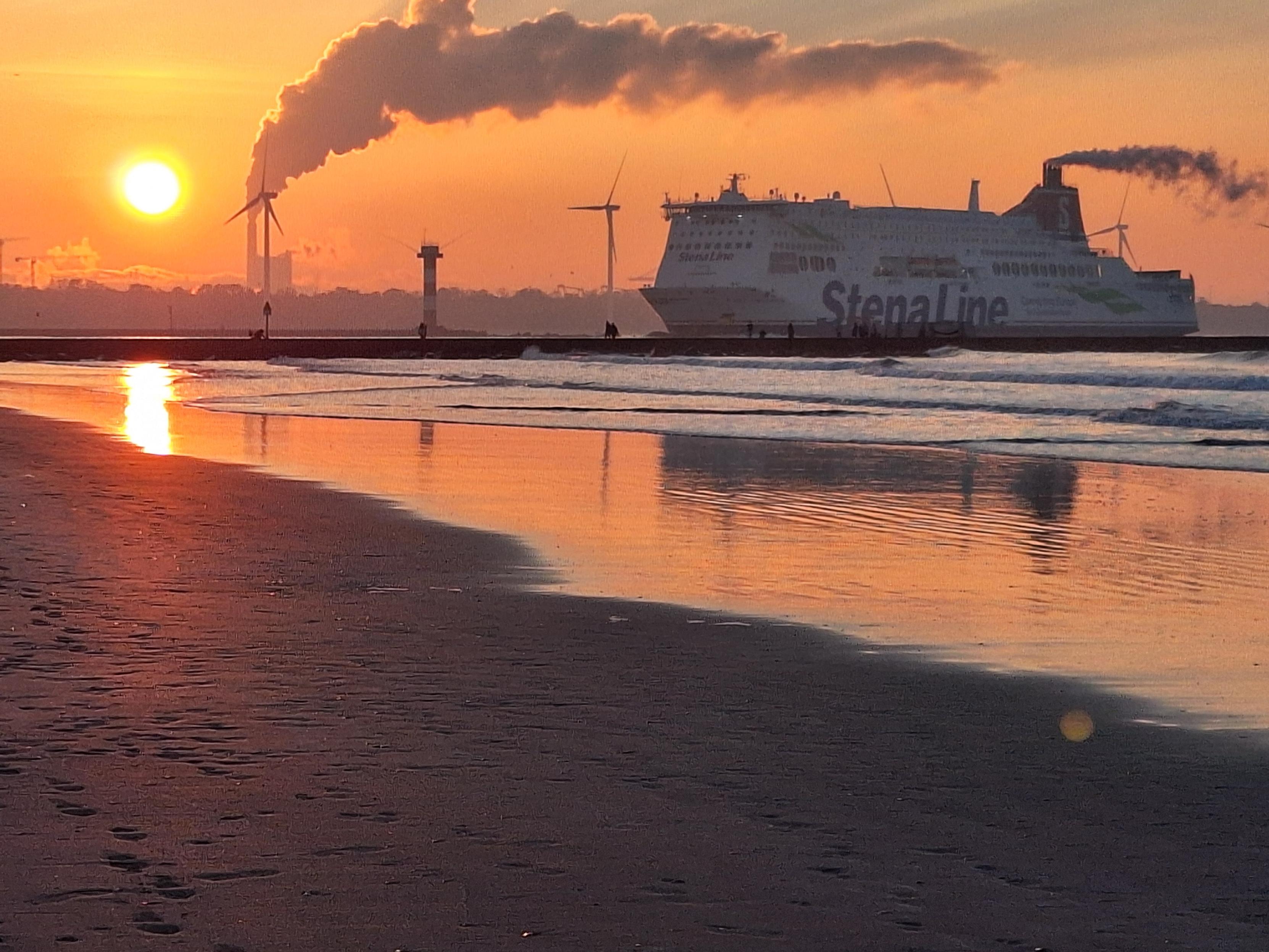 An orange sun, low in the sky. An industrial stack belching steam and fumes. A large ship emblazoned with "Stena Line" sailing from the right. In the foreground, a beach and some gently rolling waves. The orange sun and sky are reflected in the sea. Separating the ship and the beach there is a breakwater with a few people strolling.