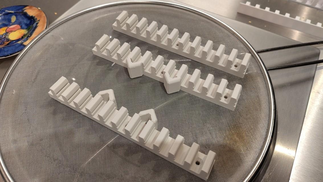3D printed supports for the grille strips. They are resting on an anti-splash screen on a wok induction hob. The rearmost is a simple castellated shape with nubs to engage with the grille strips. The front two are similar but with the addition of v-shaped prominences to accept the nuts and bolts for mounting the control panel. In the background 3D printed end caps for the grille can just be seen