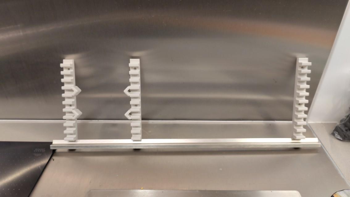 The three 3D printed slat holders with one slat attached, showing how they fit together. The slat is horizontal and the slat holders are protruding vertically above it. The two holders towards the left hand end of the slat have v-shaped receptacles for the nuts and bolts needed to hold the control panel